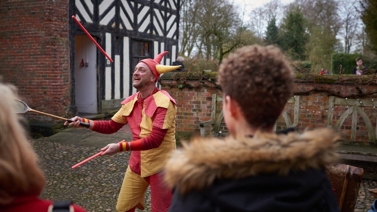 A jester dressed in yellow and red juggles to a crowd in front of a black and white Tudor house.